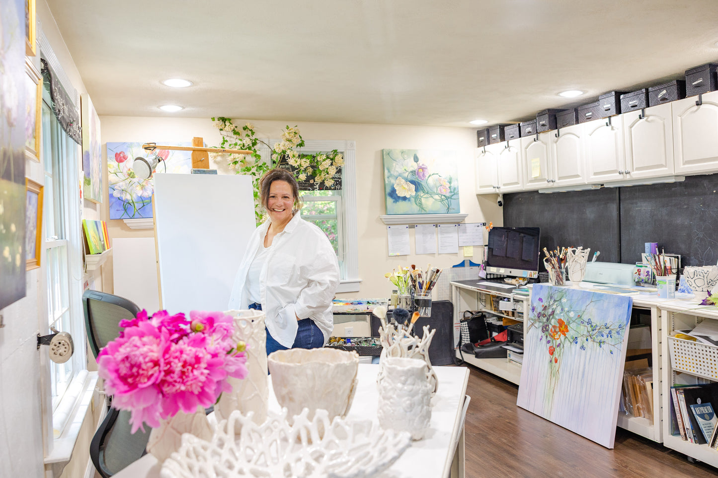 Artist Carla Thomson standing by her easel, surrounded by light and paintings in her Rhode Island studio.