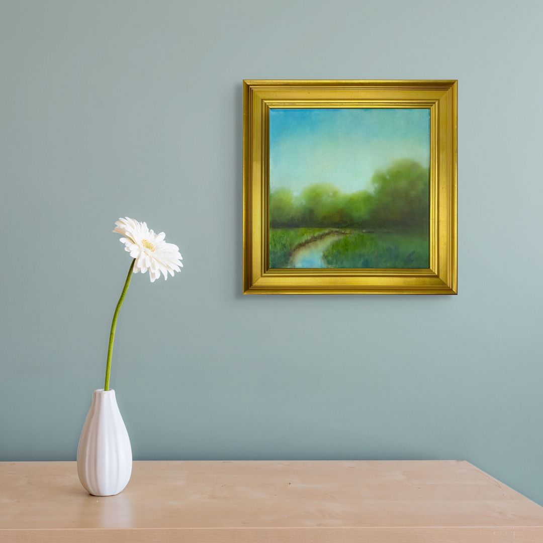 Framed original oil landscape painting on a wall above a table with a vase and flower.