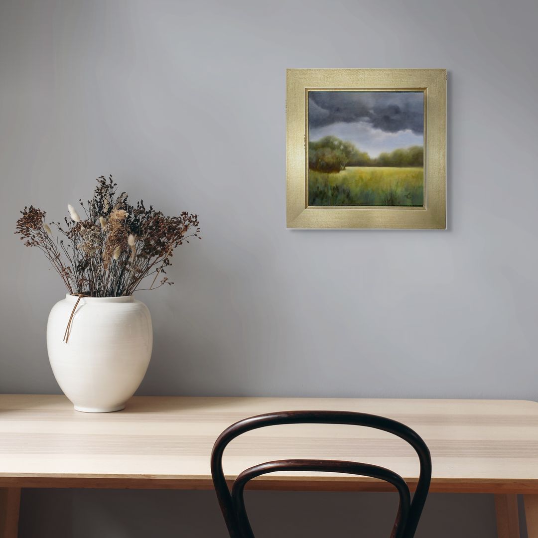 Framed original oil of green field with dark stormy sky painting on a wall above a table with a vase and chair.