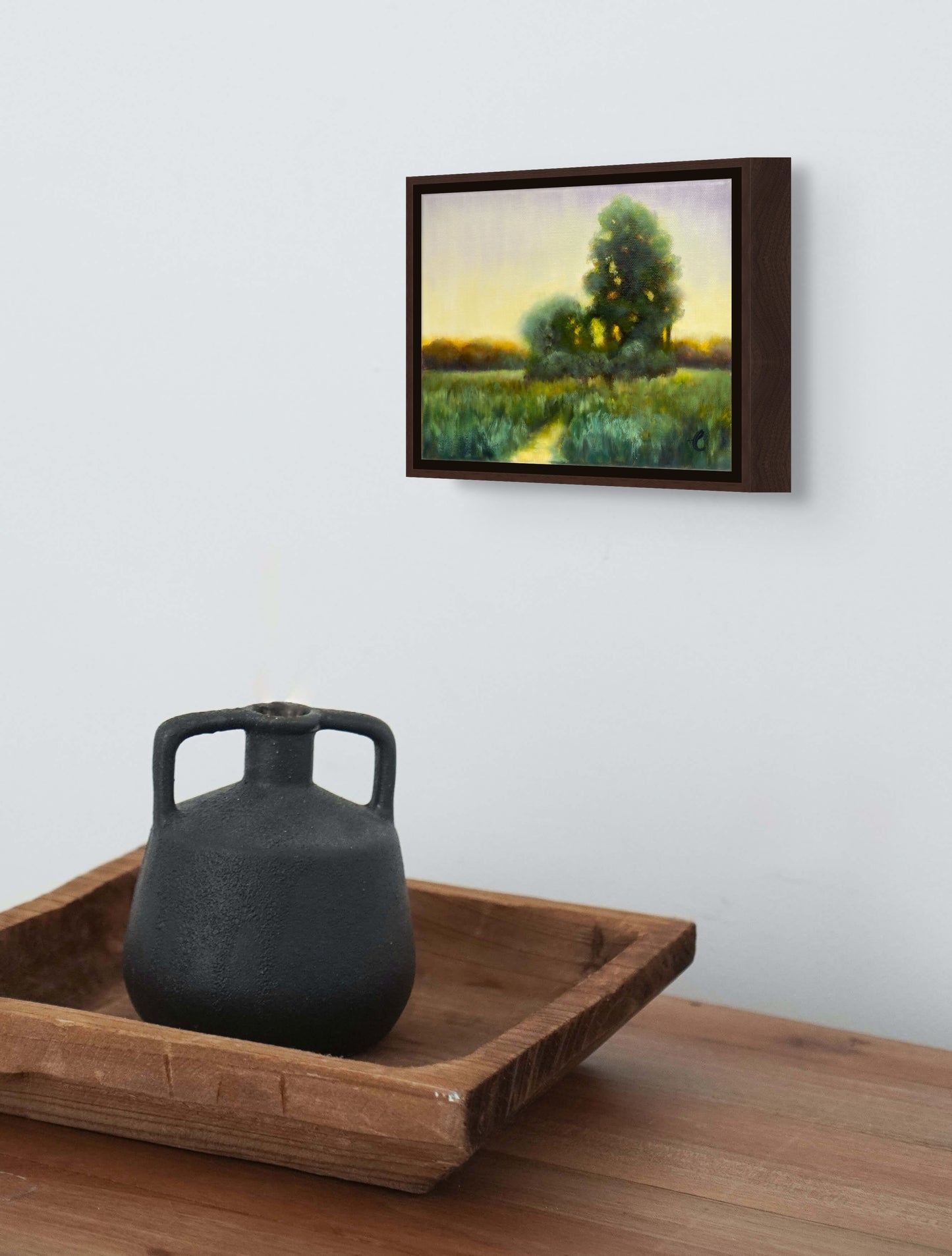 Framed painting of a landscape with a black vase on a wooden tray below.