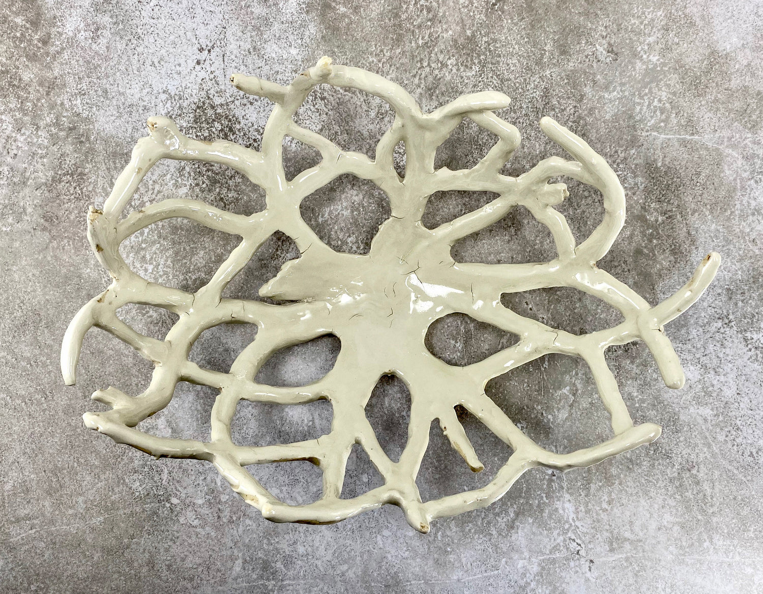 A handcrafted ceramic bowl with an openwork design resembling intertwining branches. Finished in a glossy white glaze, the sculptural form casts intricate shadows on the white surface beneath it.