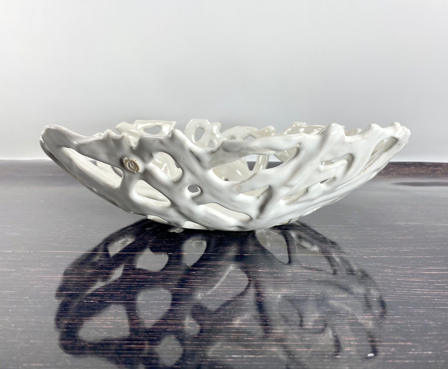 Glossy white ceramic openwork bowl with a sculptural, branch-like design, photographed on a polished dark wood table. The bowl’s intricate form is mirrored in the table’s surface, creating a dramatic reflection that highlights its elegant, hand-crafted detail.