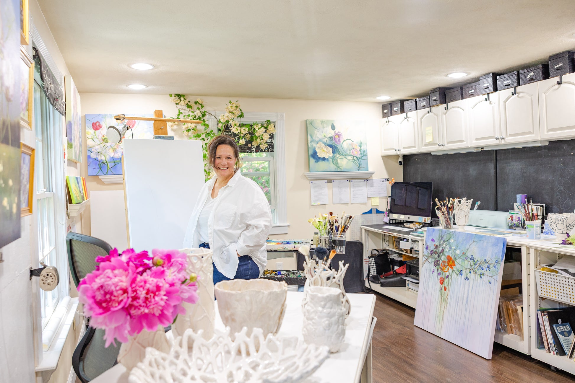 Artist Carla Thomson standing by her easel, surrounded by light and paintings in her Rhode Island studio.